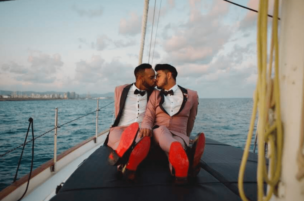 Two men in pink suits and bow ties sitting on a boat, leaning close and about to kiss, with a city skyline and cloudy sky in the background.