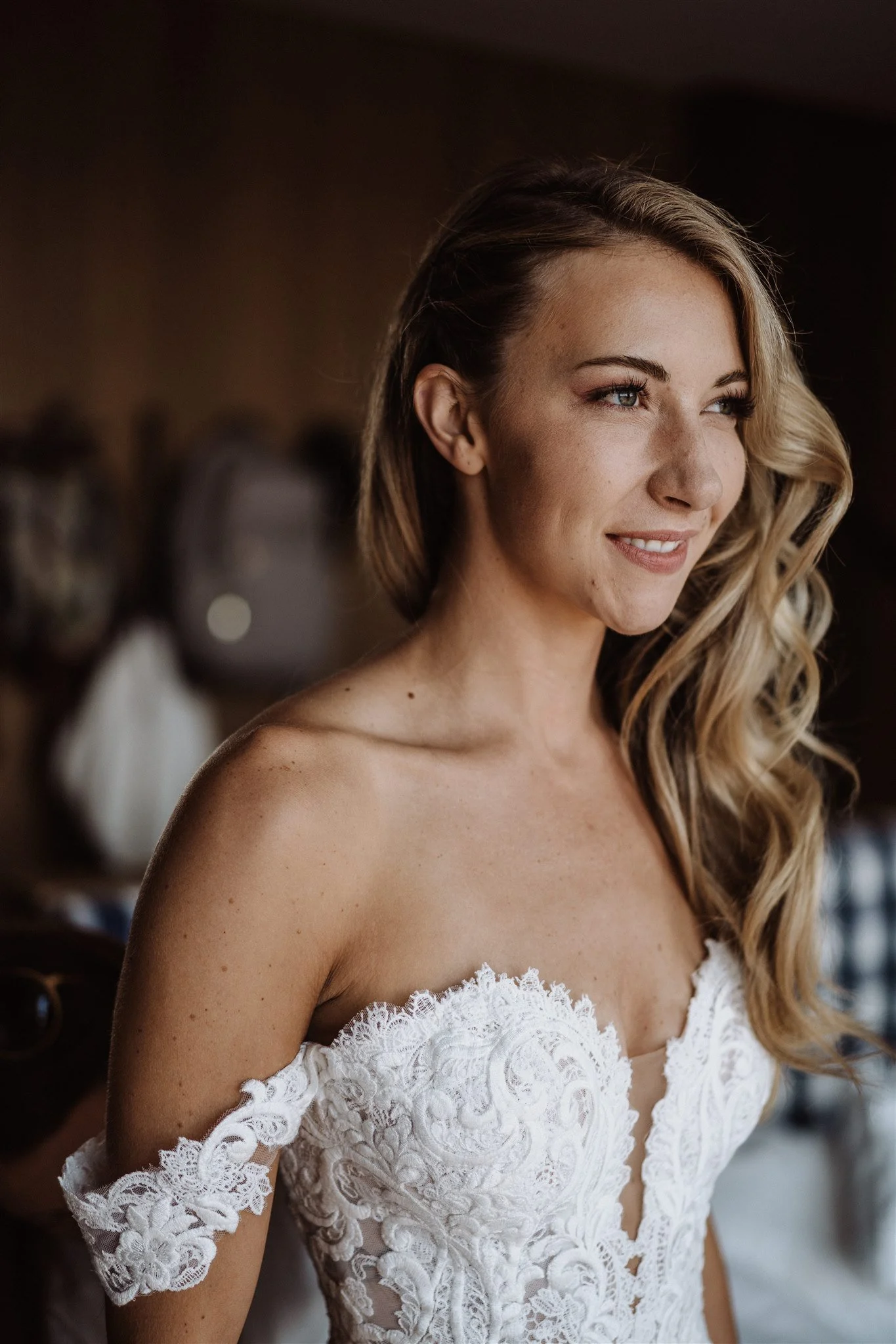 A woman with wavy blonde hair and blue eyes wearing a white lace off-shoulder dress, smiling softly, indoors with a dark background.