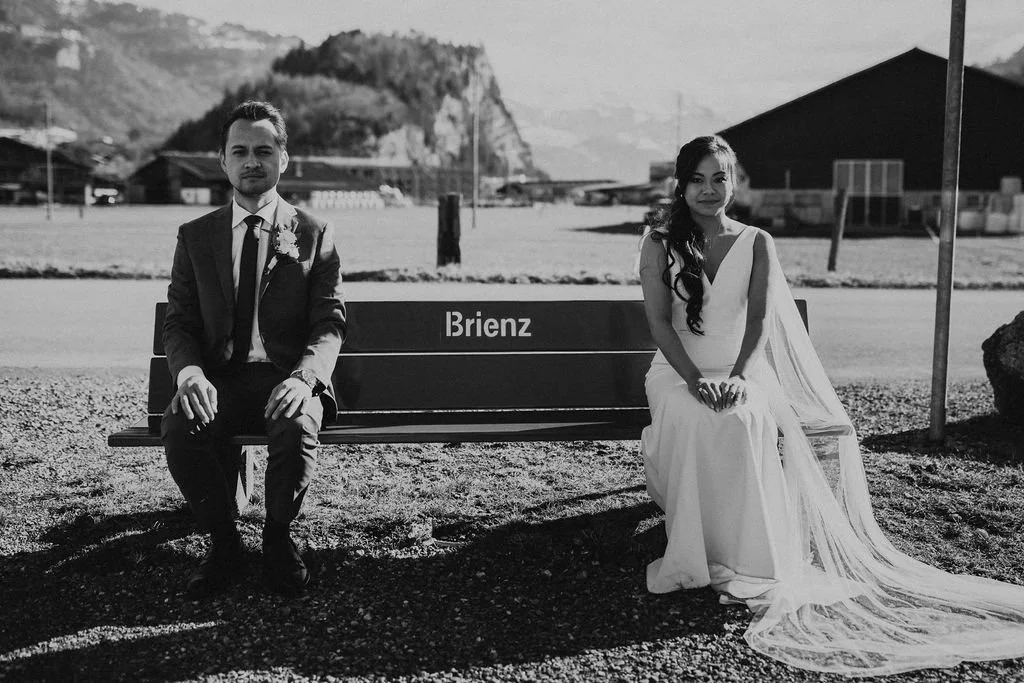 A couple in wedding attire sitting on a park bench labeled "Brienz" outdoors with mountains in the background in black and white.