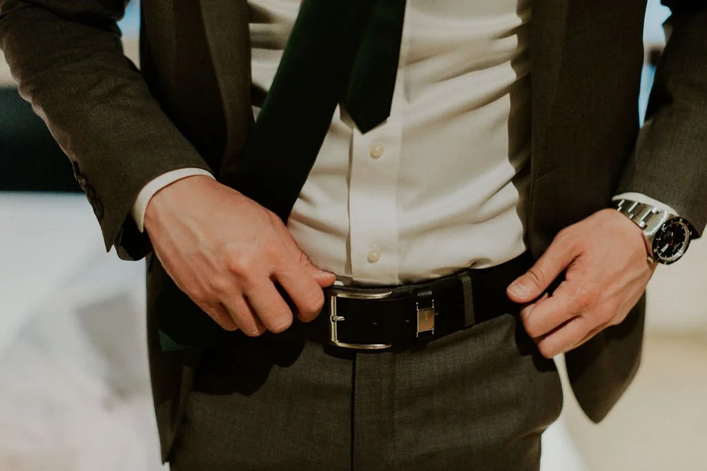 Close-up of a man in a gray suit with a white shirt and black tie, adjusting his black belt with a silver buckle.