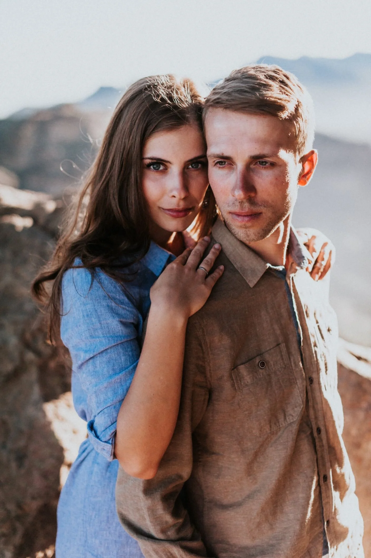 A young woman with long dark hair and blue eyes leaning close to a young man with short blonde hair and blue eyes, outdoors with mountains in the background.