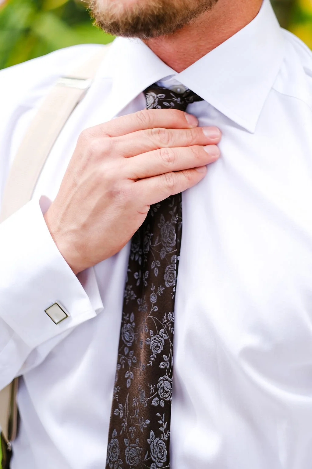 A man adjusting his black floral tie with his right hand, wearing a white dress shirt with French cuffs and a silver square cufflink, outdoors with greenery in the background.