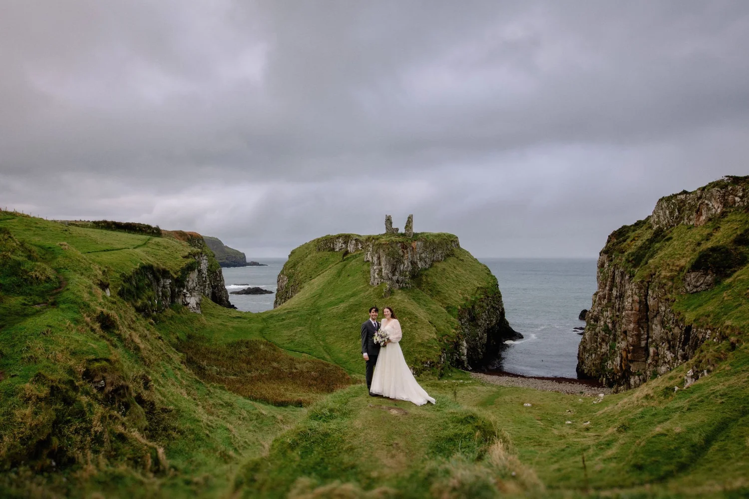 A bride and groom standing on a grassy hillside near the coast with cliffs and ocean in the background, under cloudy skies.
