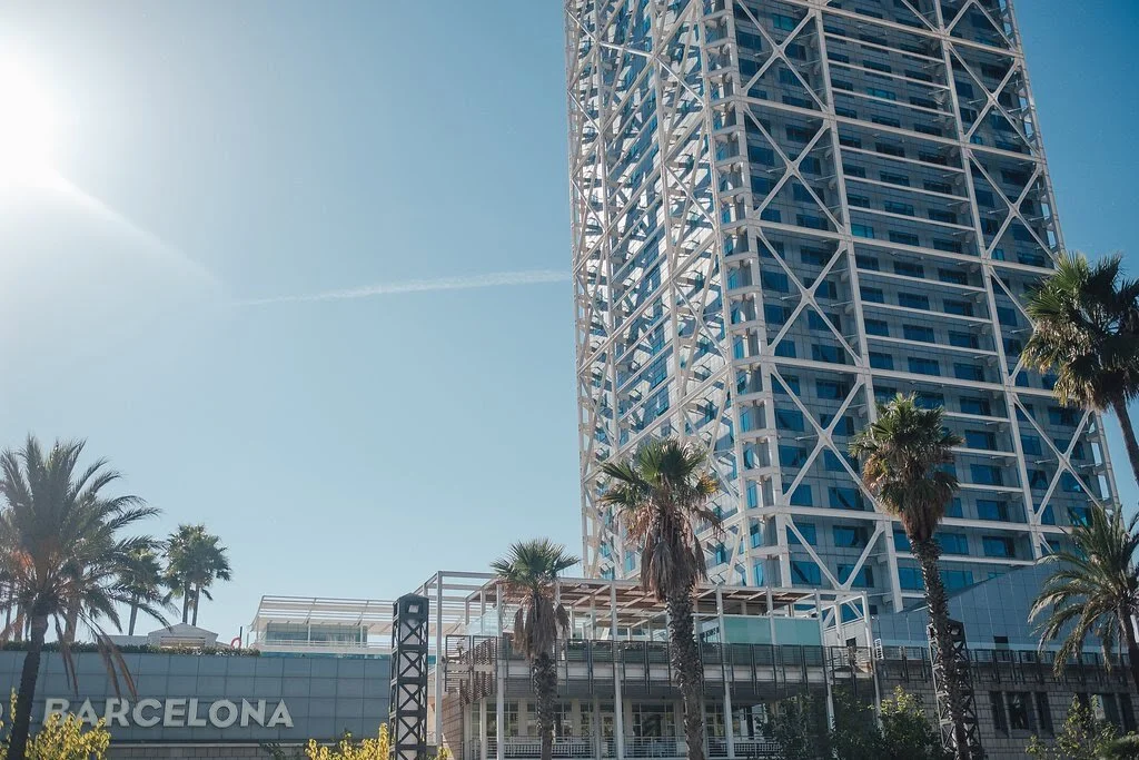 Tall modern skyscraper with white lattice framework and glass windows, palm trees in front, clear blue sky, and a sign reading "Barcelona" at the bottom left.