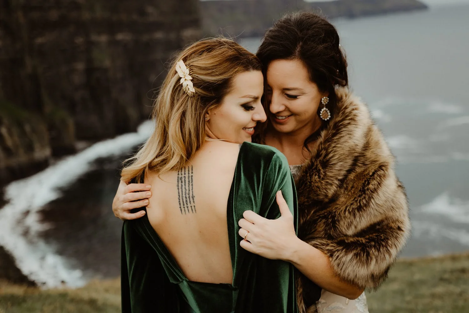 Two women hugging outdoors by water with cliffs in background, one with blonde hair and a leaf hair accessory, the other with dark hair and large earrings, both smiling