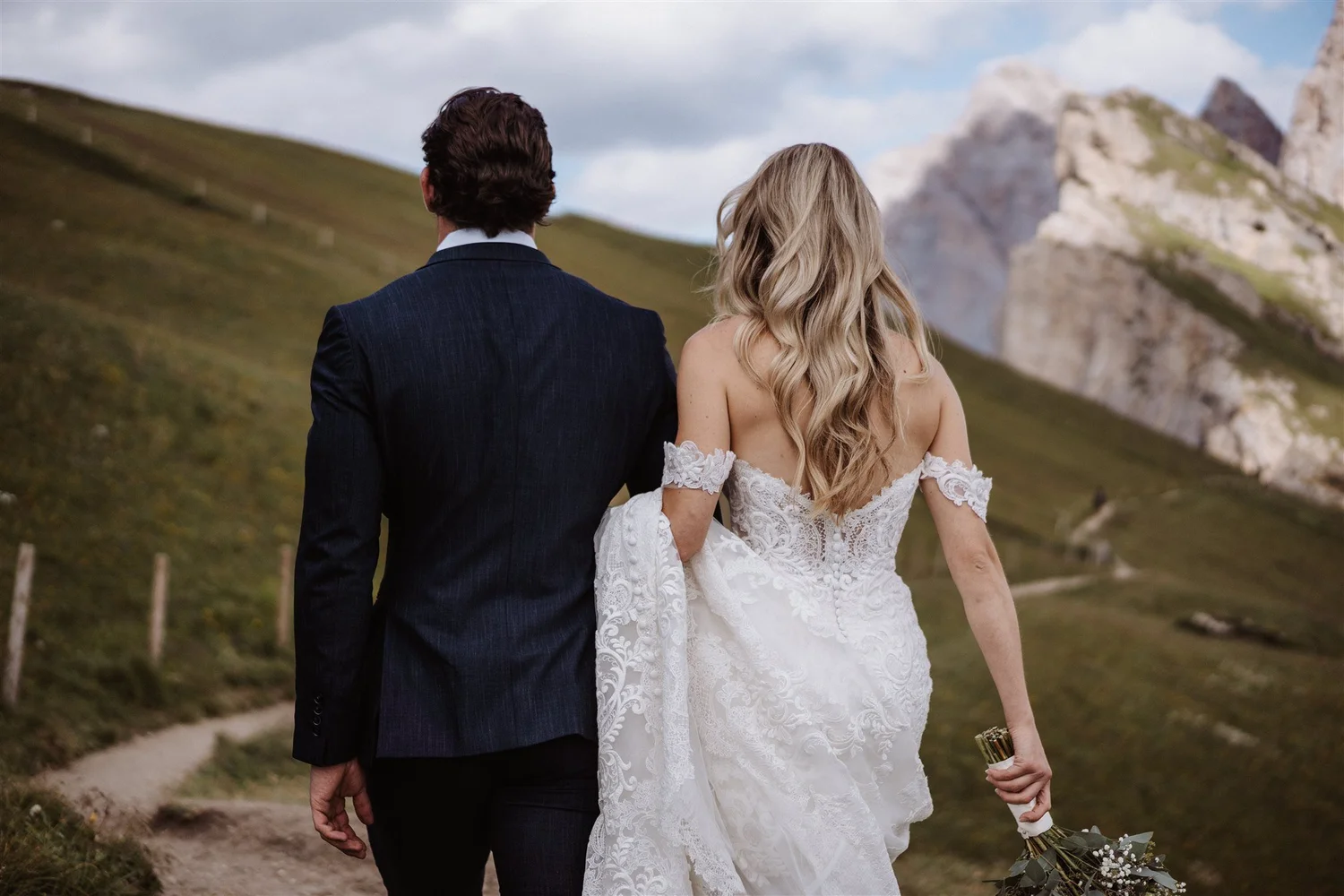 A bride and groom walking away in a mountainous landscape, the bride holding a bouquet and wearing a white lace wedding dress, the groom in a dark suit.
