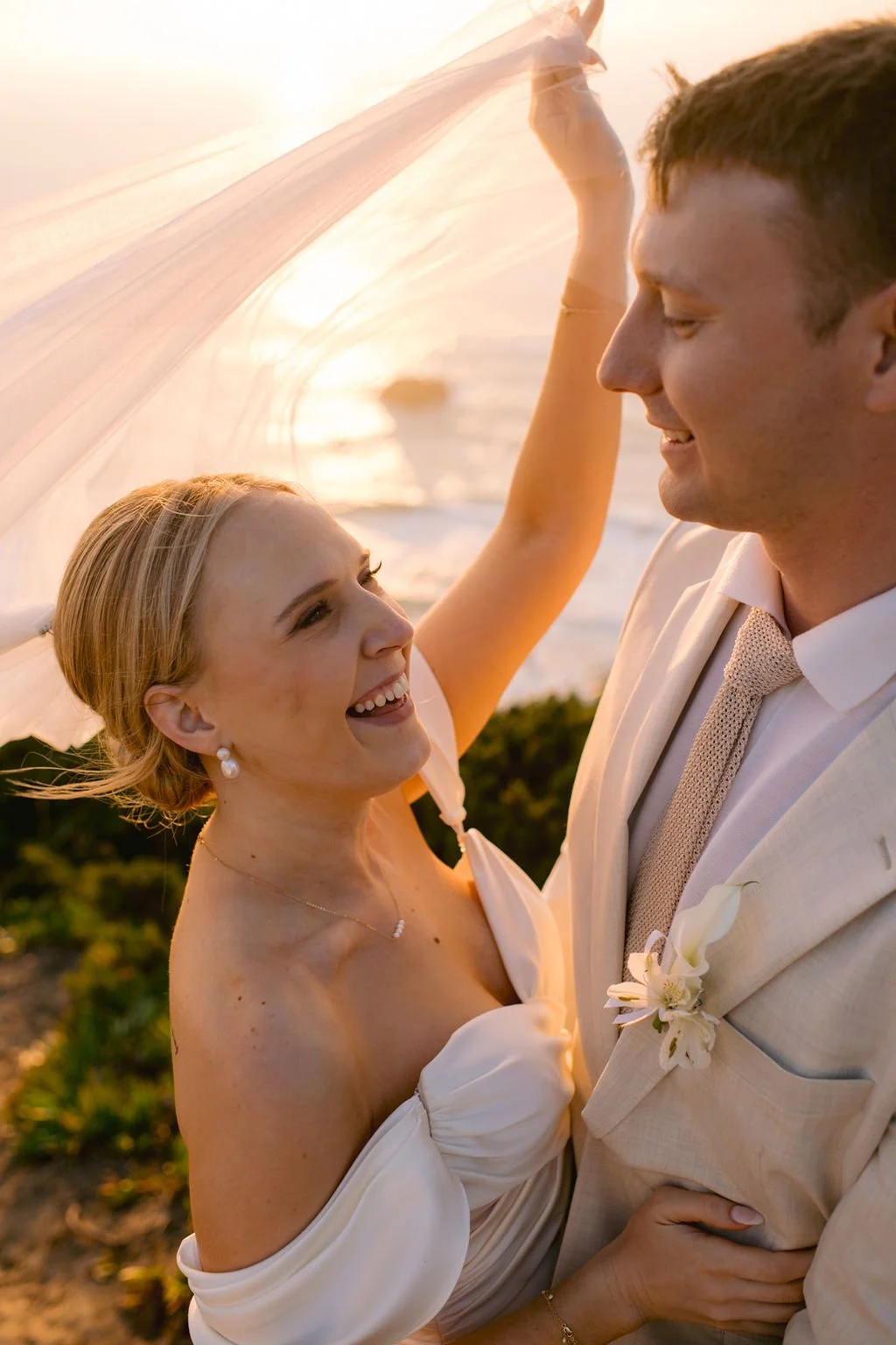 A couple in wedding attire smiling and looking into each other's eyes on a beach during sunset, with the bride lifting a sheer veil.