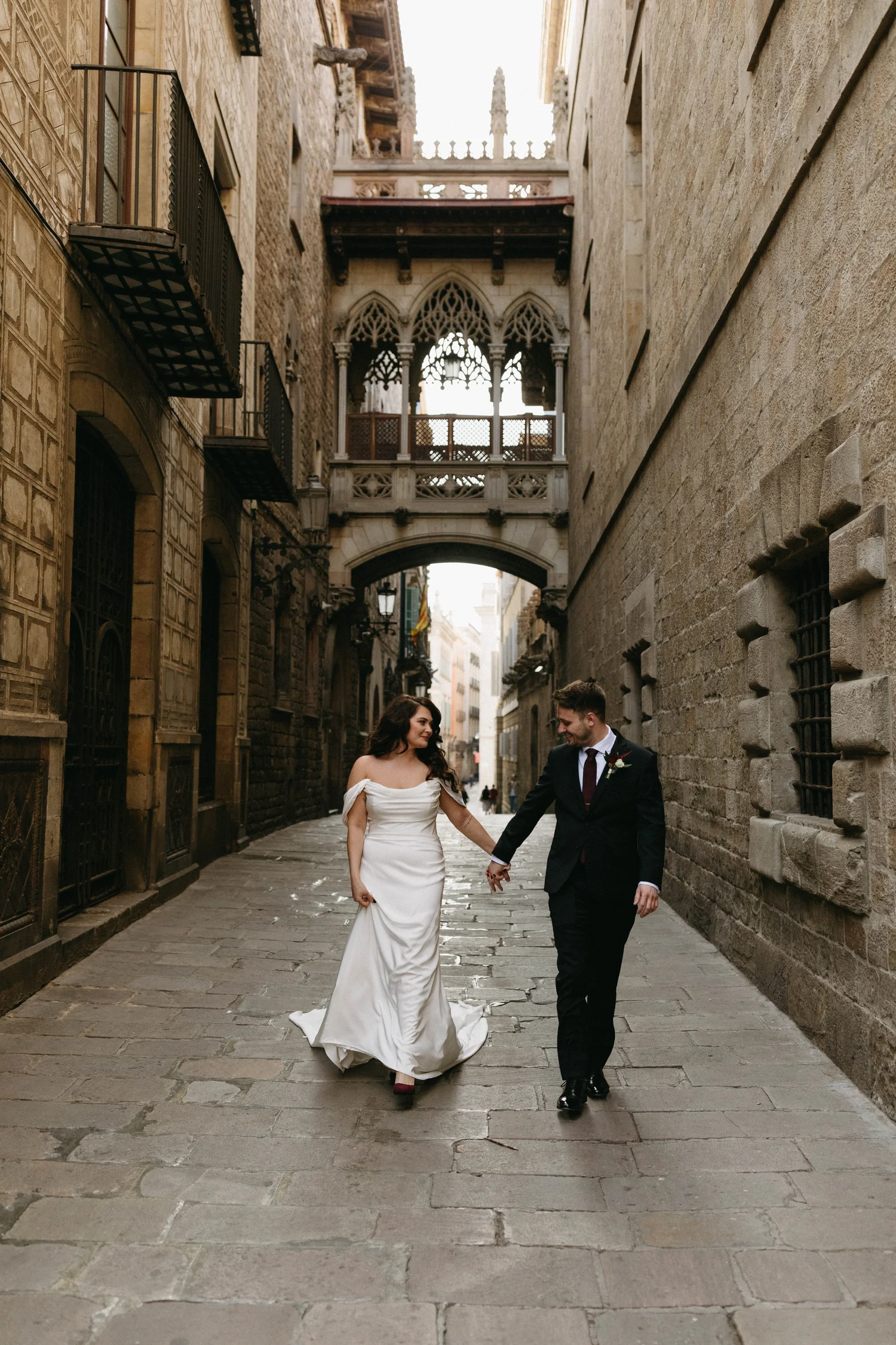 A bride in a white wedding dress and a groom in a black suit holding hands and walking down a narrow stone street in an old European city.