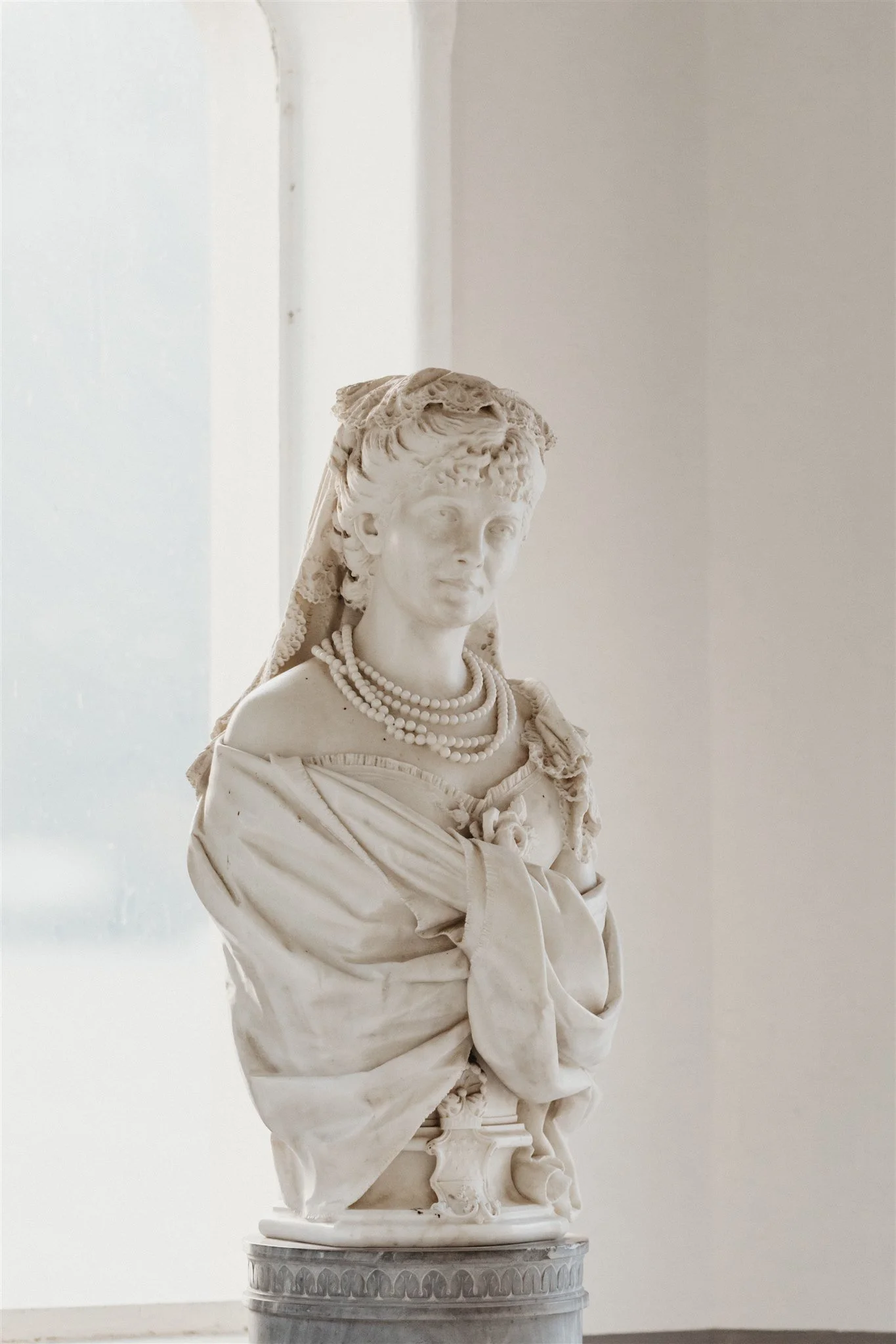 Marble bust of a woman wearing pearl necklaces and a lace head covering, displayed on a pedestal.
