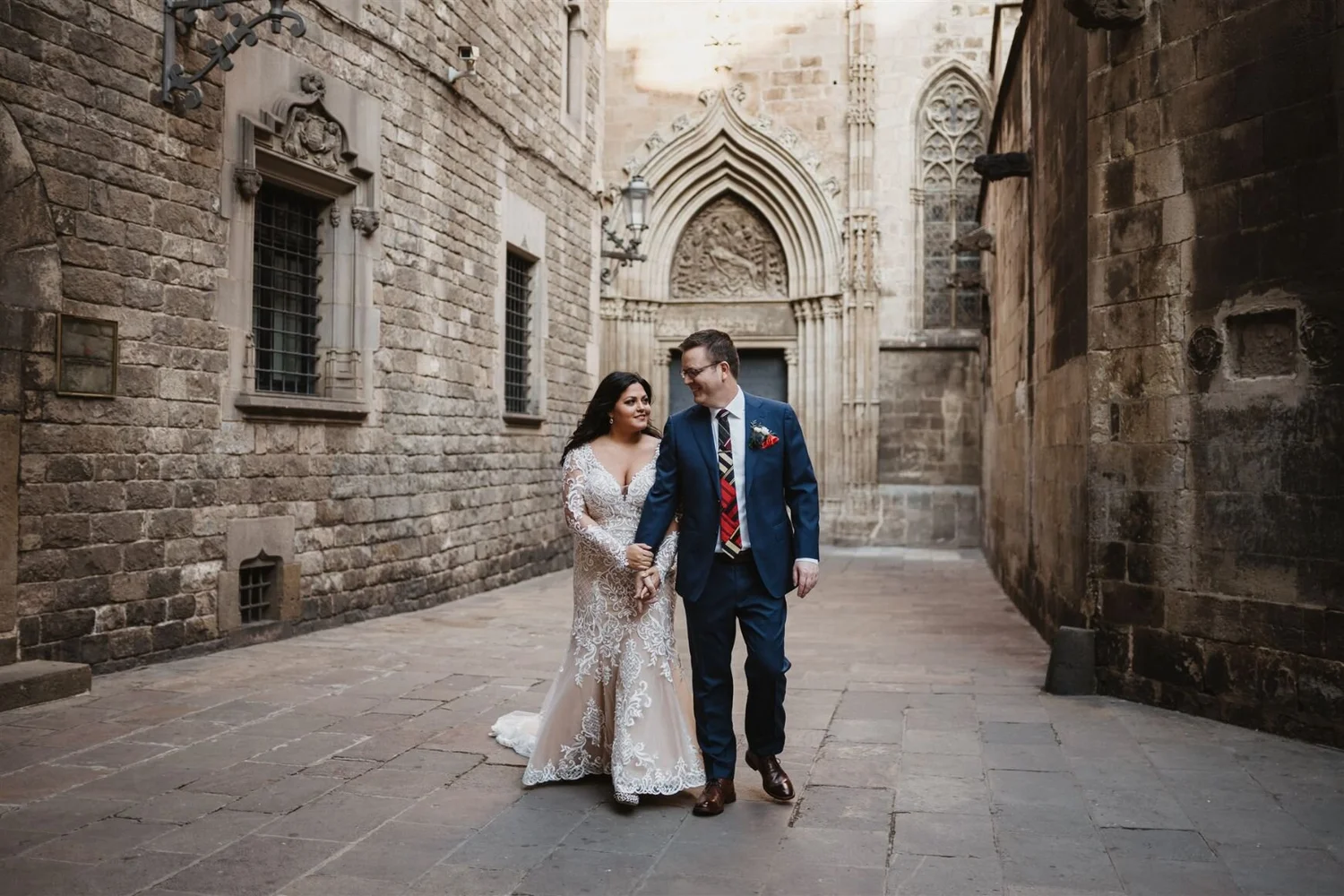 A newlywed couple walking hand-in-hand through a historic stone alleyway, with the bride in a lace wedding gown and the groom in a navy suit, surrounded by old stone buildings and gothic architectural details.