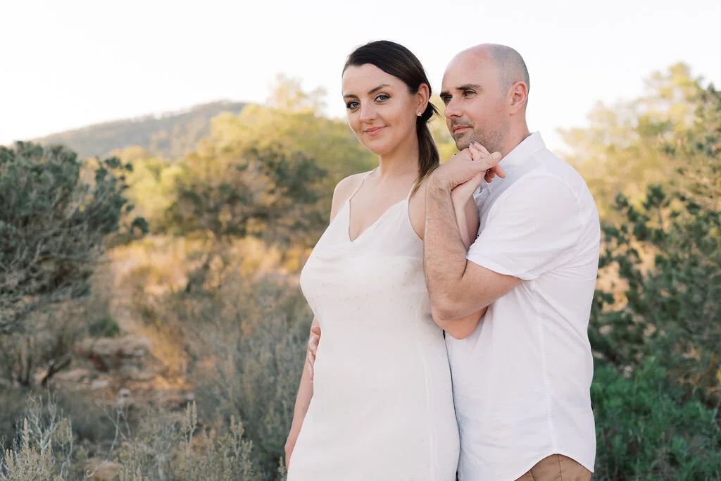 A man and woman standing outdoors, the woman with dark hair and wearing a white dress, the man with a shaved head and wearing a white shirt, in a natural landscape with trees and hills in the background.