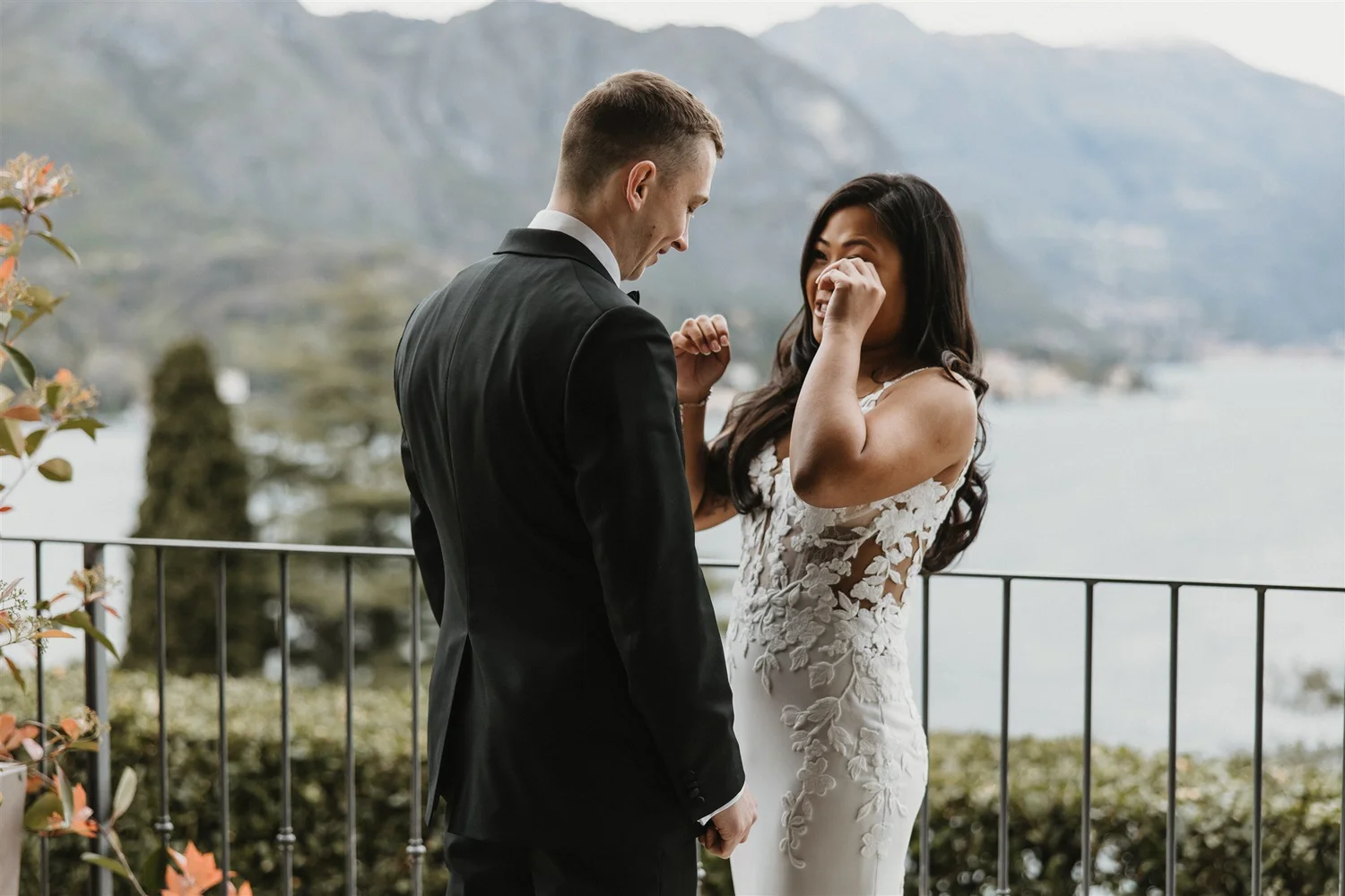 A couple dressed in wedding attire standing outdoors on a balcony, with a scenic mountain and lake view in the background. The man is in a tuxedo, and the woman is in a white wedding dress, both sharing an emotional moment.