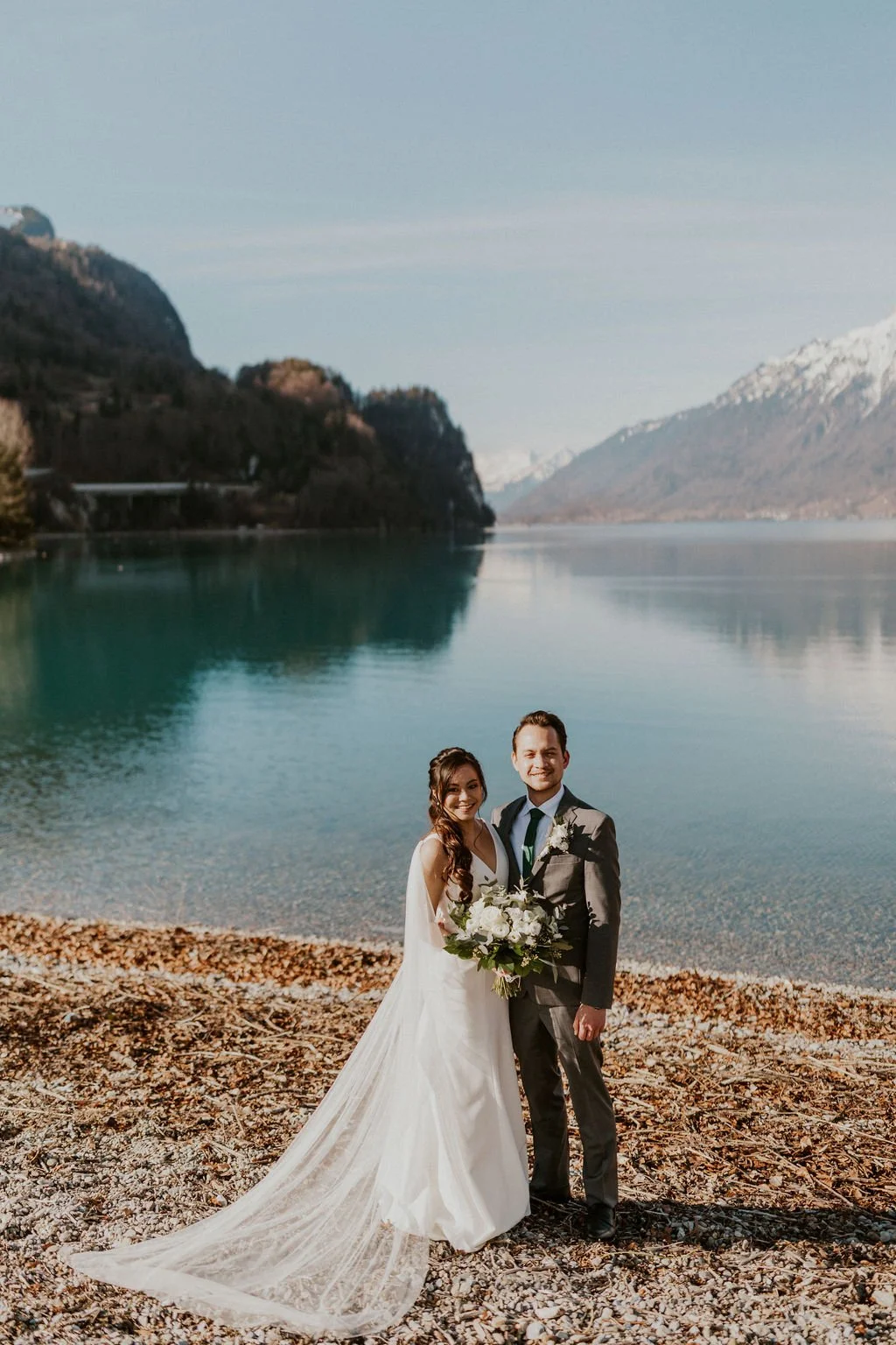 Bride and groom standing by a lake with mountains in the background, smiling on their wedding day.