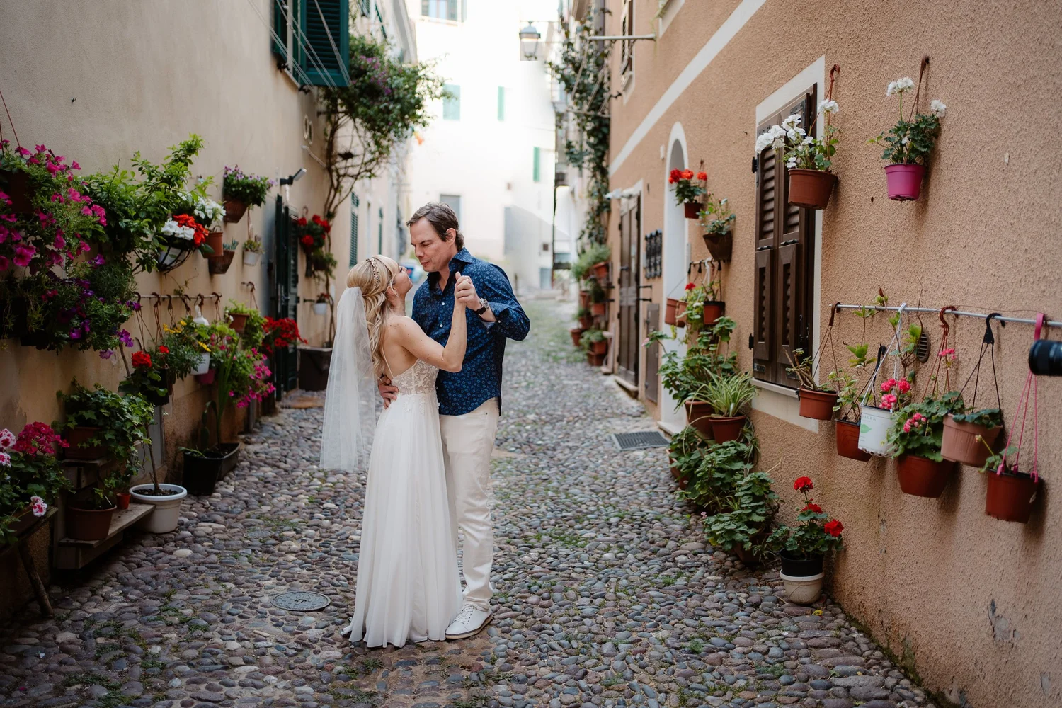 A couple dancing in an alleyway with cobblestone pavement, surrounded by potted plants and flowers on the walls.