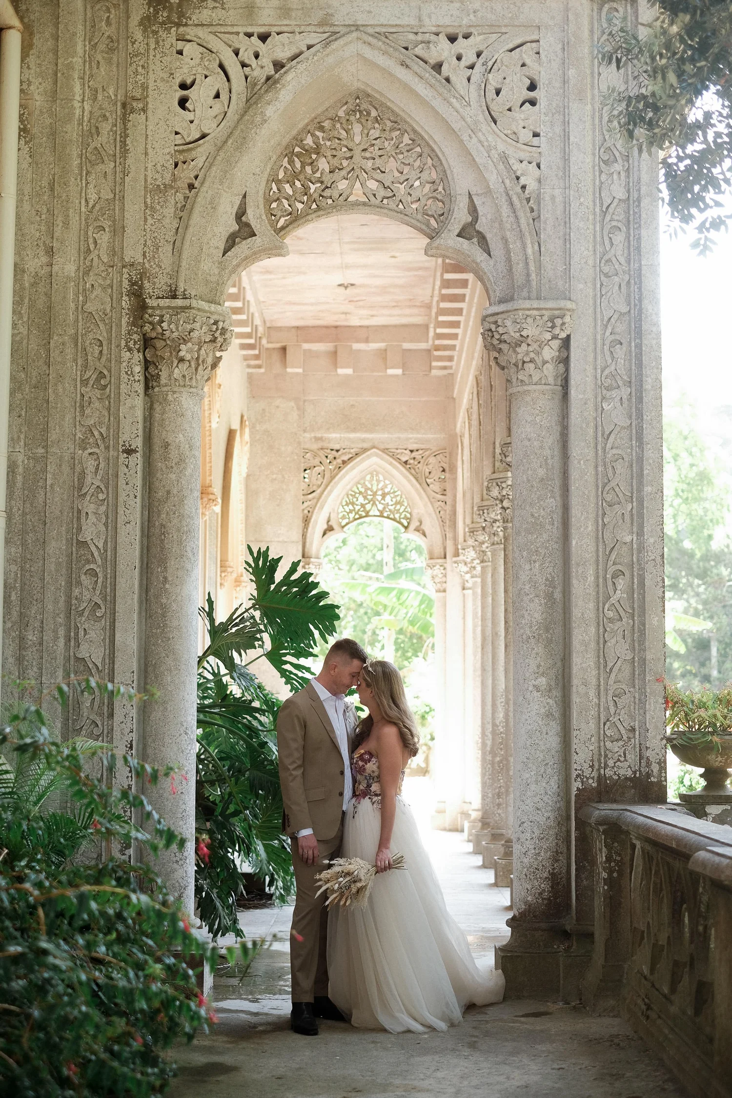 A bride and groom standing closely together, touching foreheads, in an elegant, ornate hallway with stone arches and lush green plants.