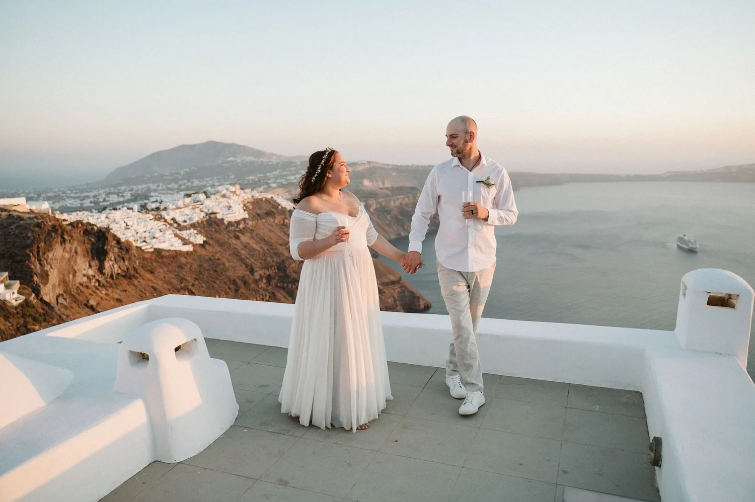 A bride and groom holding hands and celebrating on a balcony overlooking the ocean and cliffs at sunset, with a cruise ship in the water.