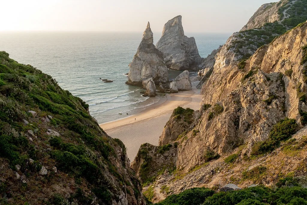 A scenic view of a beach with large rock formations in the ocean, seen through rocky cliffs covered in green vegetation.
