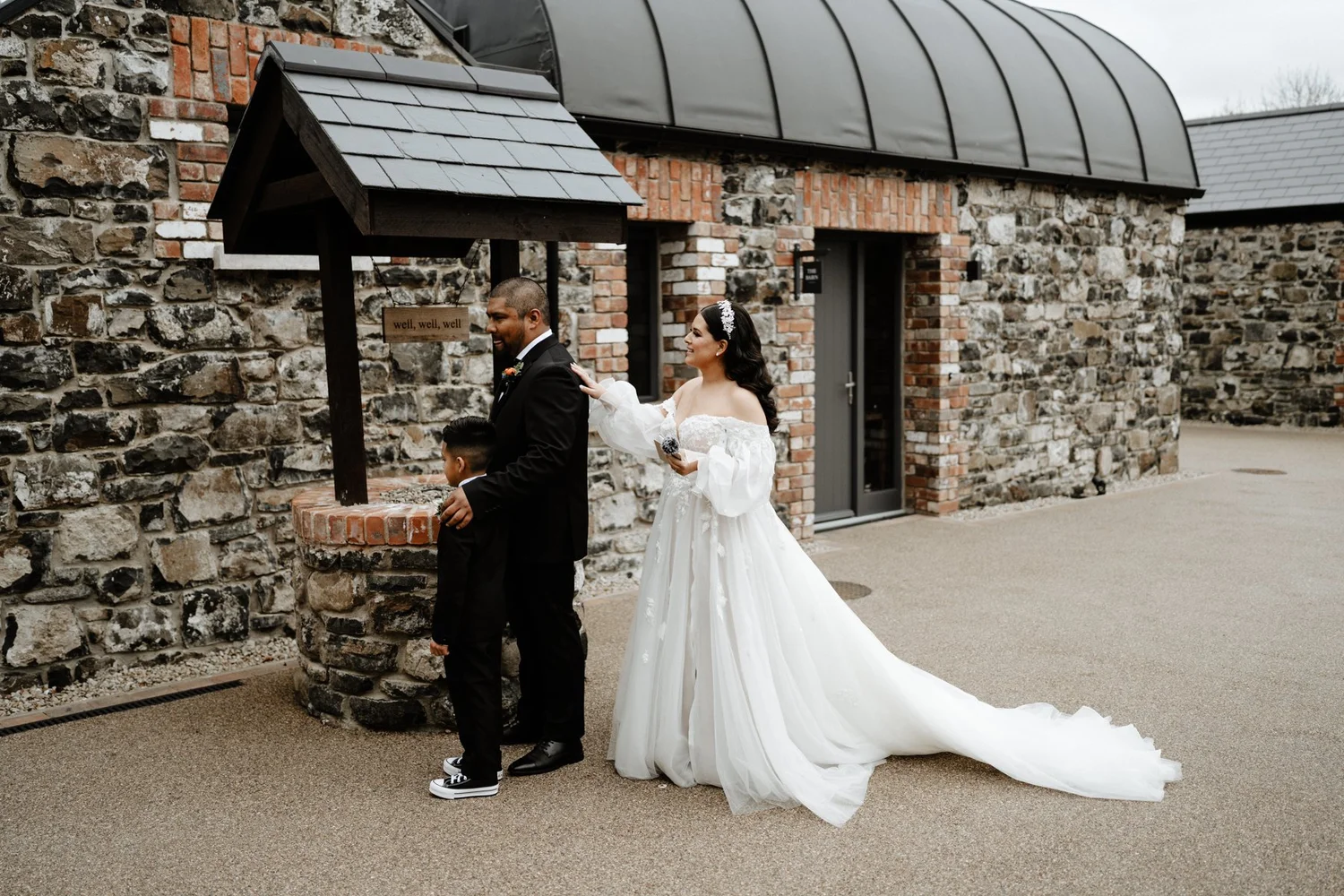 A bride in a white wedding gown with lace details and a veil standing outside a stone building, with a man in a black tuxedo and a young boy in a black suit. The man and boy are facing away from the bride near a sign with the words "well, well, well"