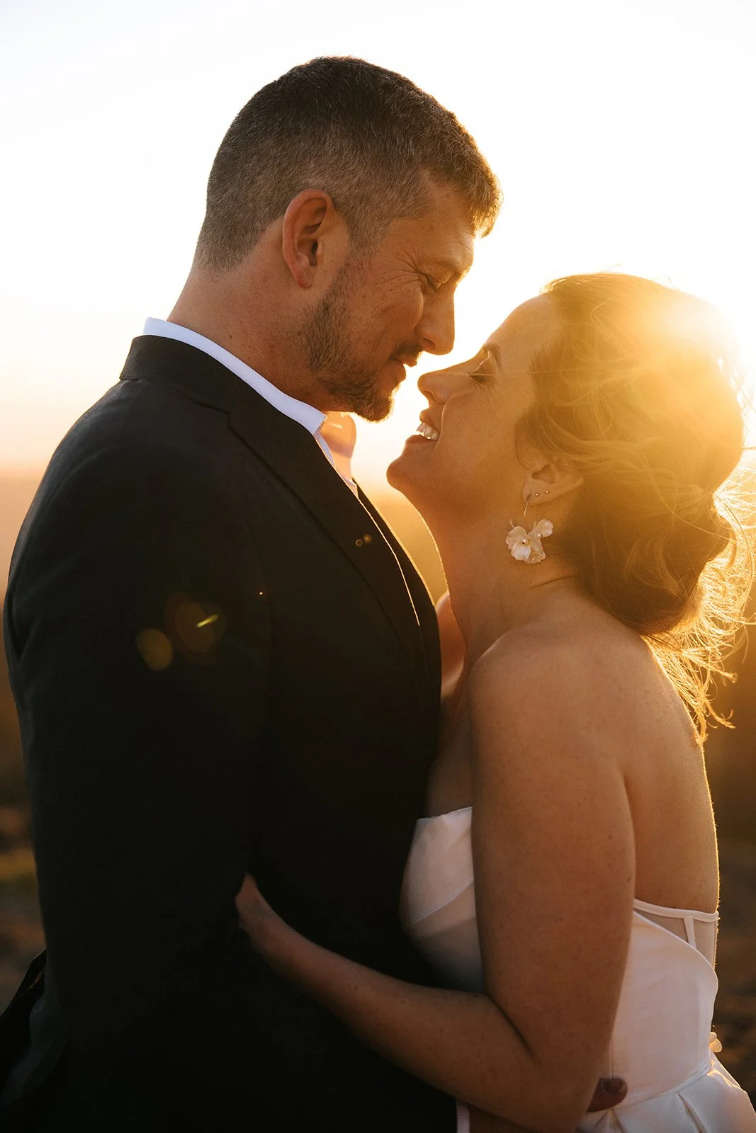 A couple dressed in wedding attire, embracing and smiling at each other during sunset.